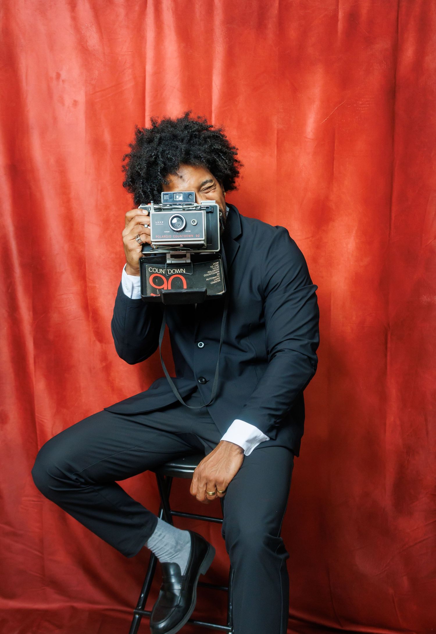 Photographer in a black suit strikes dynamic poses while holding vintage cameras against a vibrant red backdrop. Birmingham photographer. A