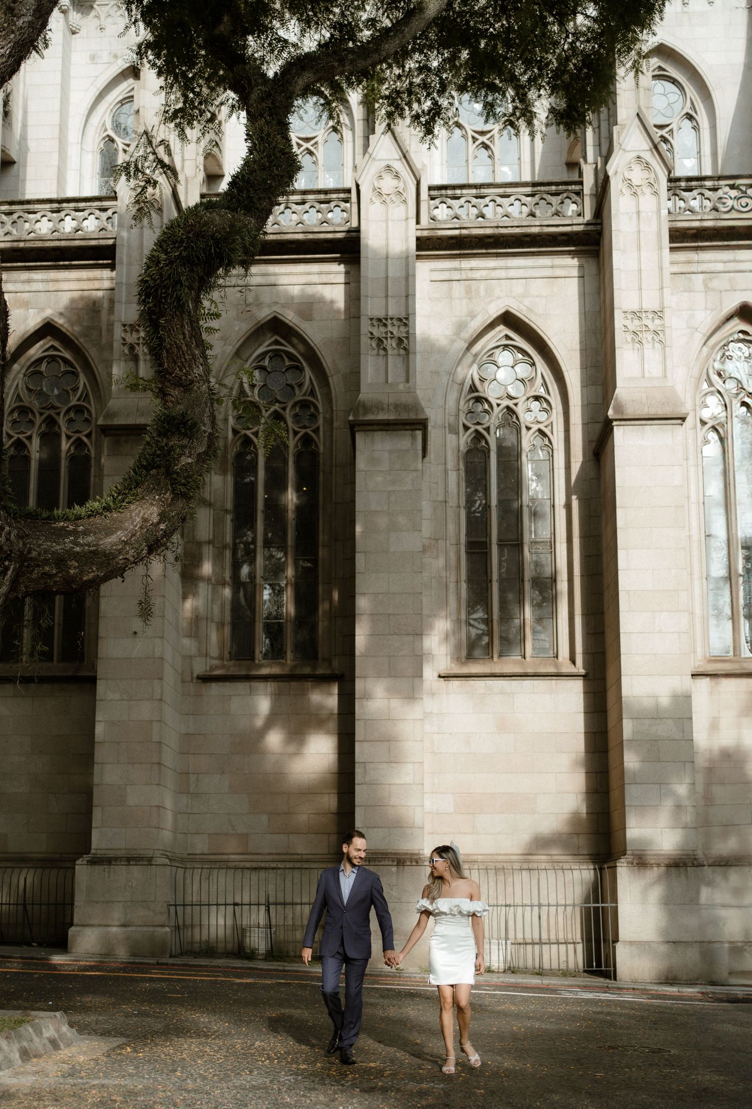 Couple walking beside gothic church architecture with overhanging tree branches.
