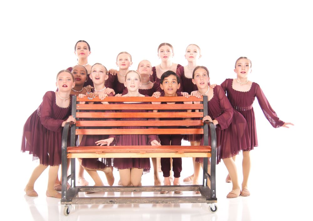 Young dancers in burgundy dresses pose playfully around a wooden bench in a studio setting.