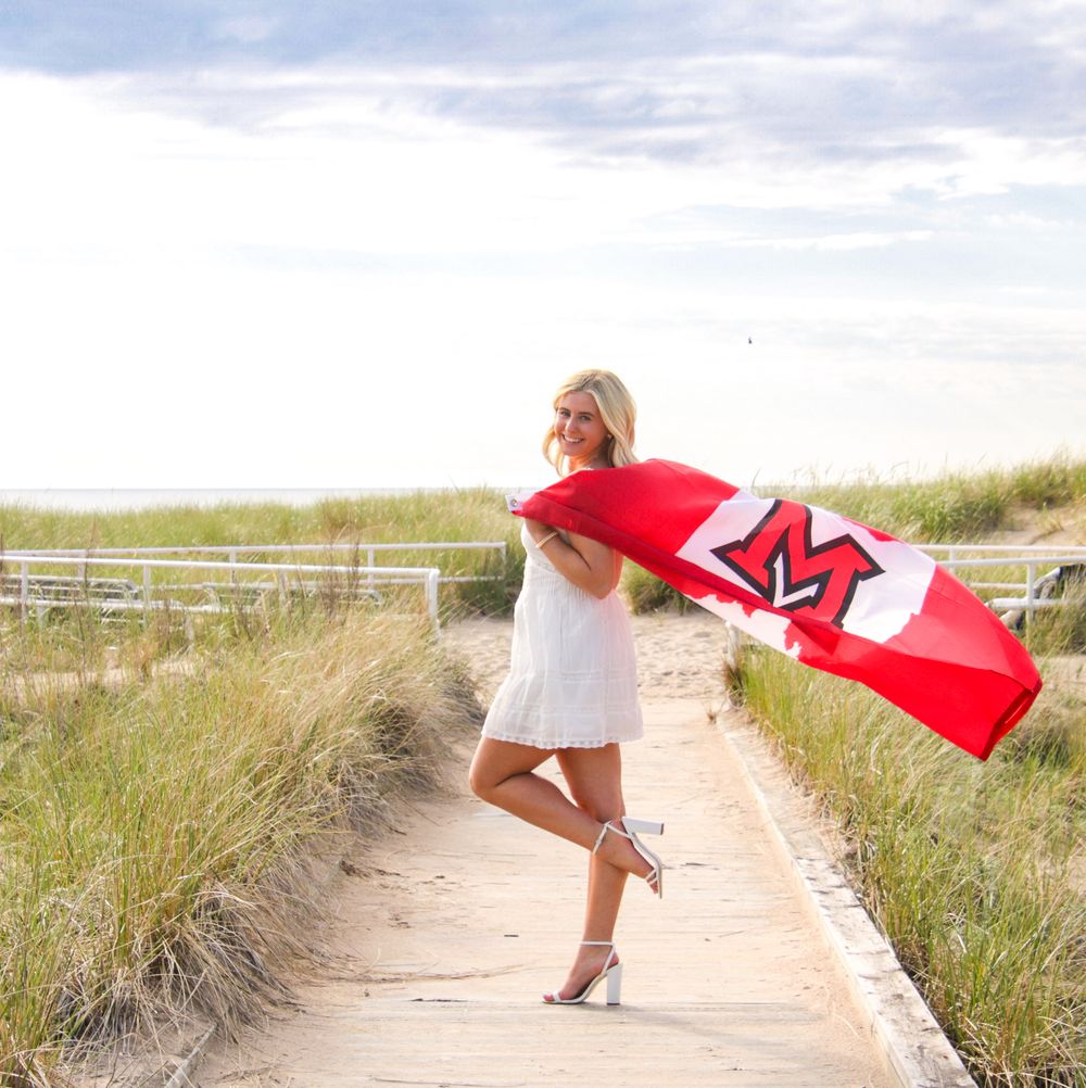 A figure in a white dress holds a red surfboard on a sandy beach path.