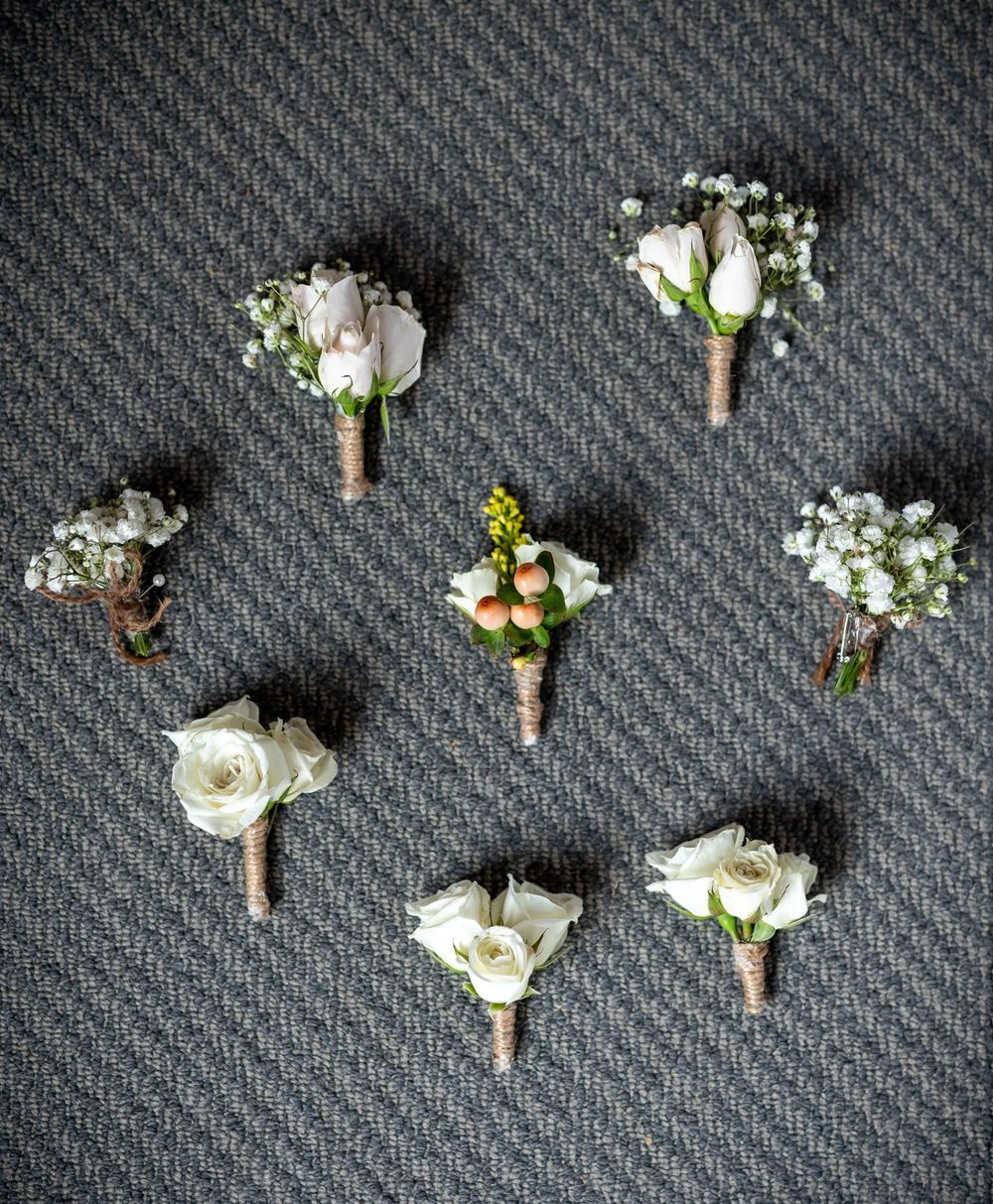 Collection of white rose boutonnieres with baby's breath arranged on grey textured fabric.