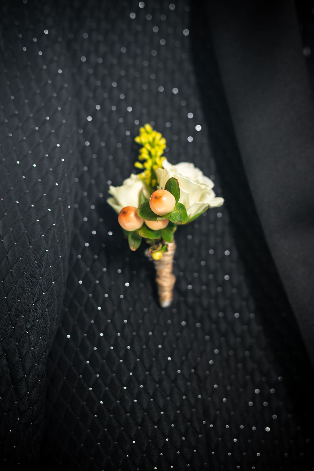 A delicate boutonniere with white roses and yellow flowers on a textured black fabric.