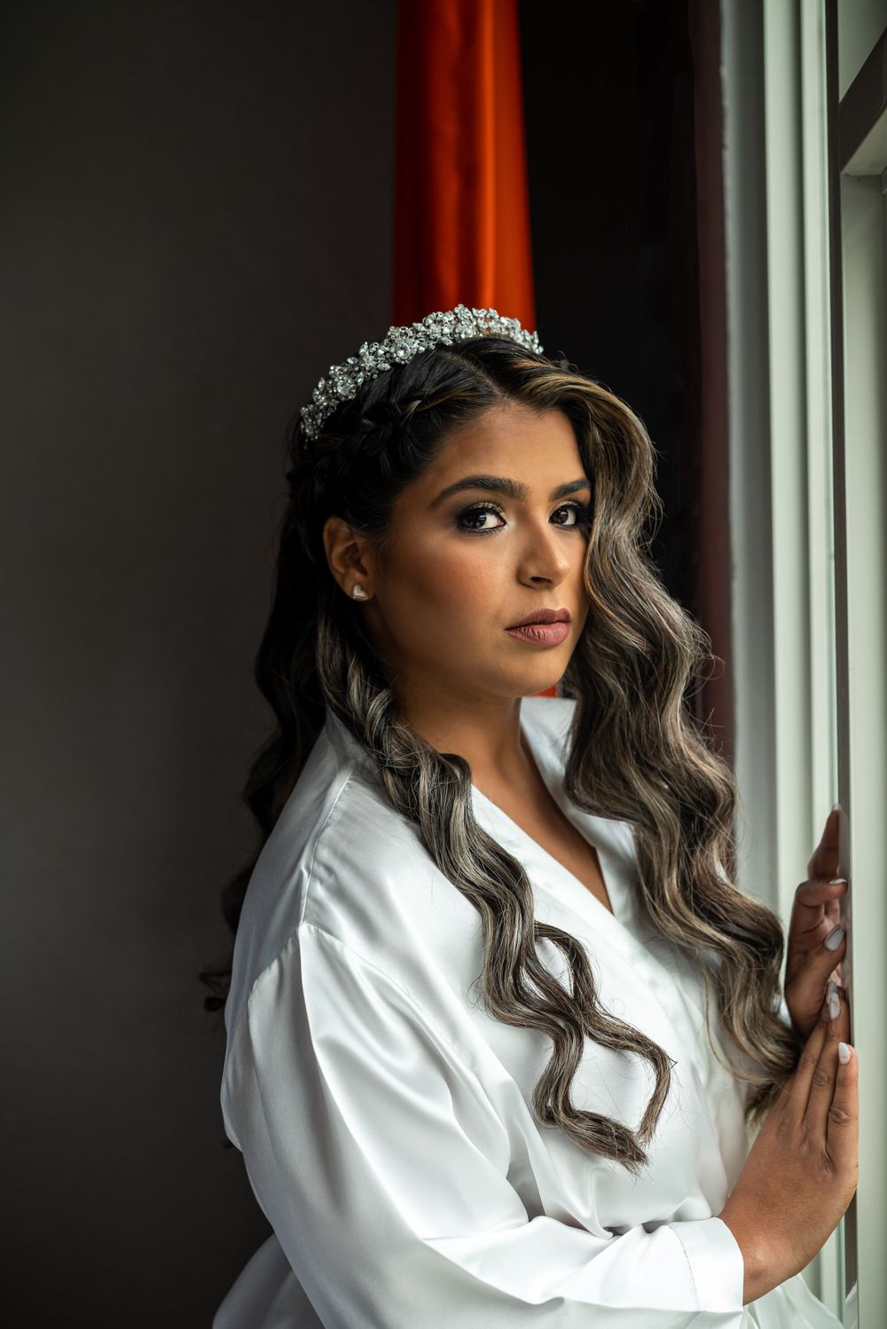 Bride in white robe and crystal crown poses by window with dramatic lighting.