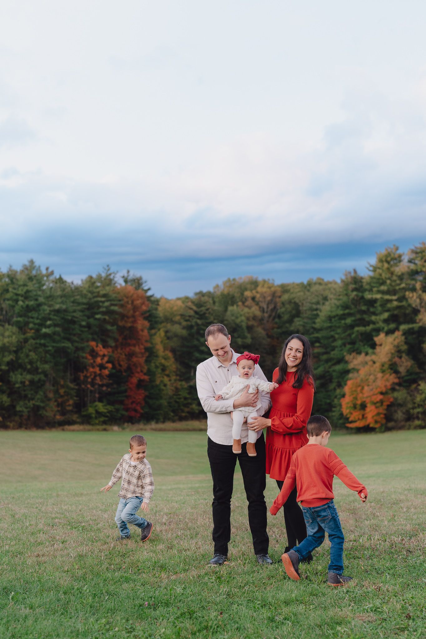 Family enjoys a playful autumn moment in an open field surrounded by colorful fall foliage under cloudy skies.