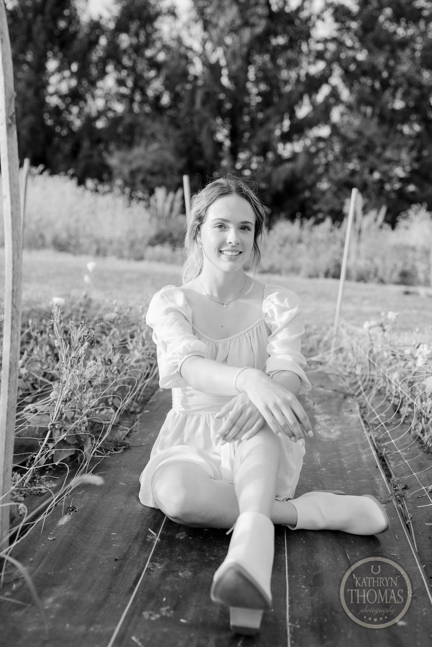 A black and white portrait shows someone in a light dress seated on dark planks outdoors.
