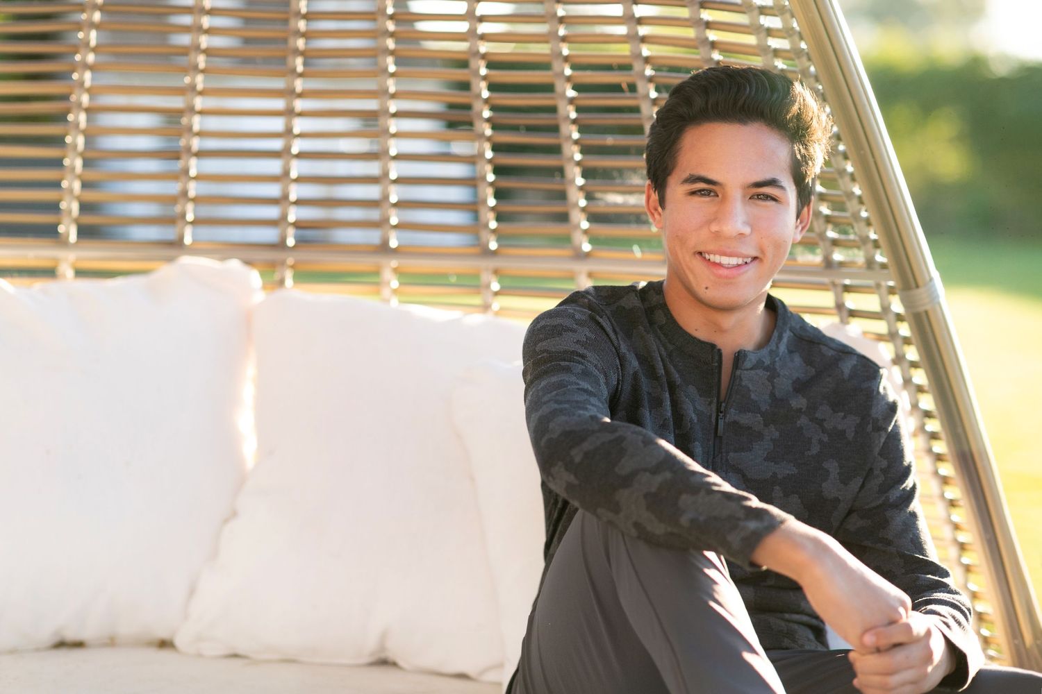 A high school senior relaxes on an outdoor hanging chair wearing a dark sweater on a sunny day.