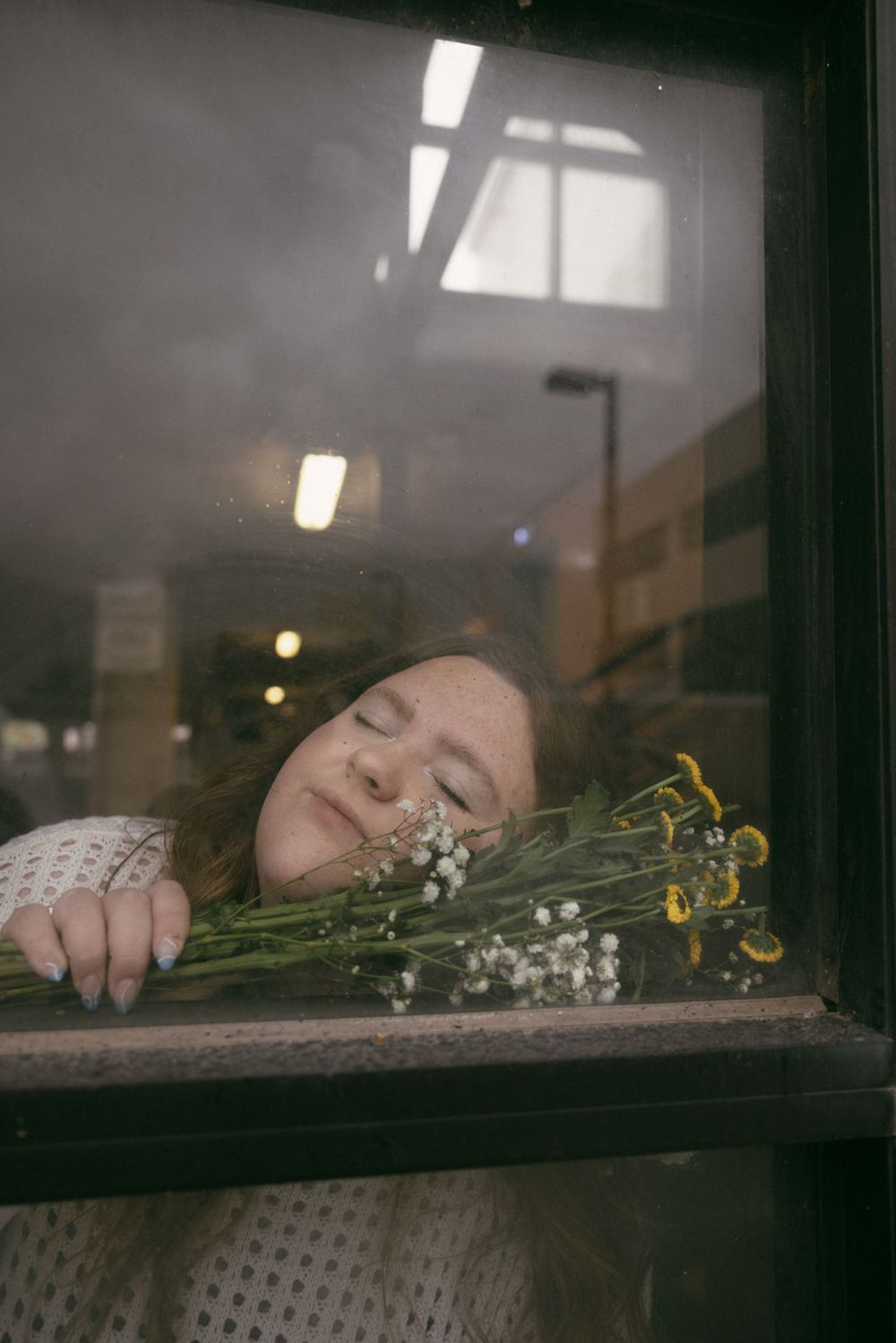 Dreamy artistic photo through window glass showing someone resting with delicate flowers in a moody setting.