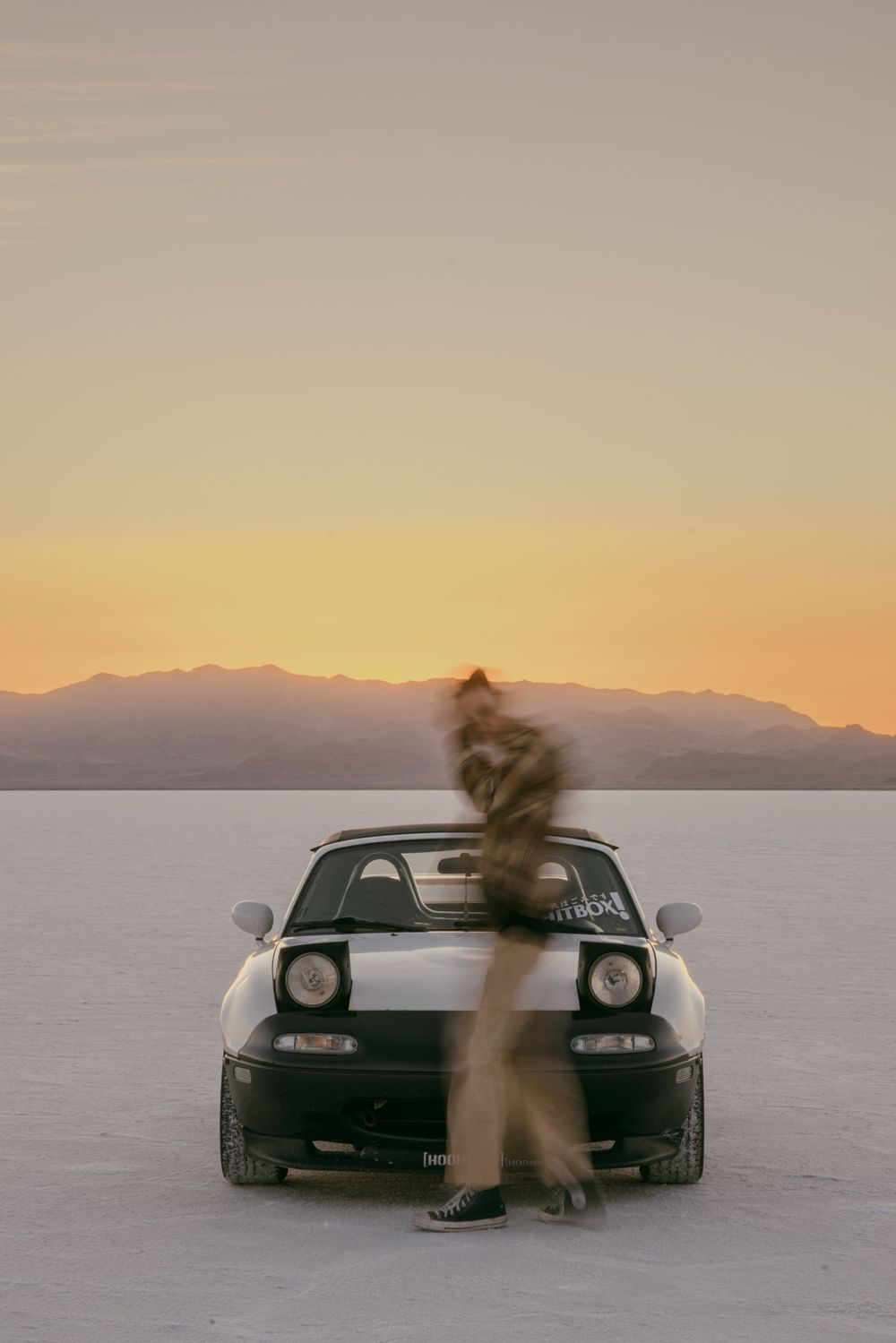 Silhouetted figure standing in front of sports car on salt flats during sunset.