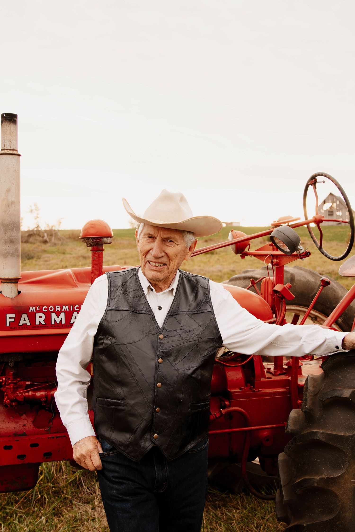 A person in western attire stands proudly next to a classic red Farmall tractor in a rustic farm setting.