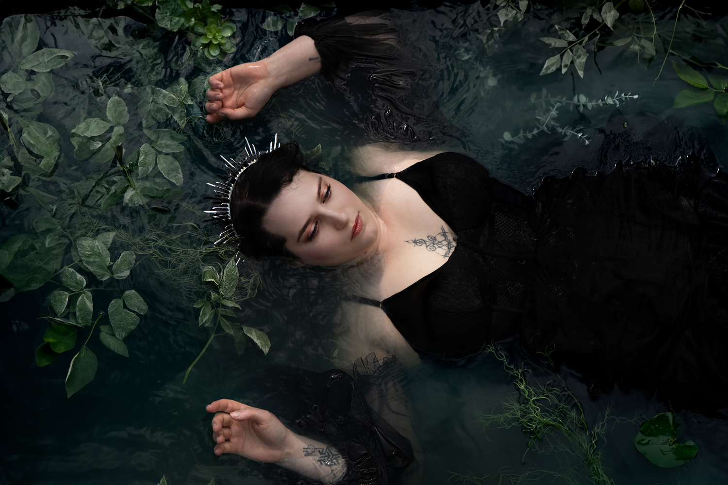 Goth crowned woman submerged in a pond like Ophelia. lily pads surround her