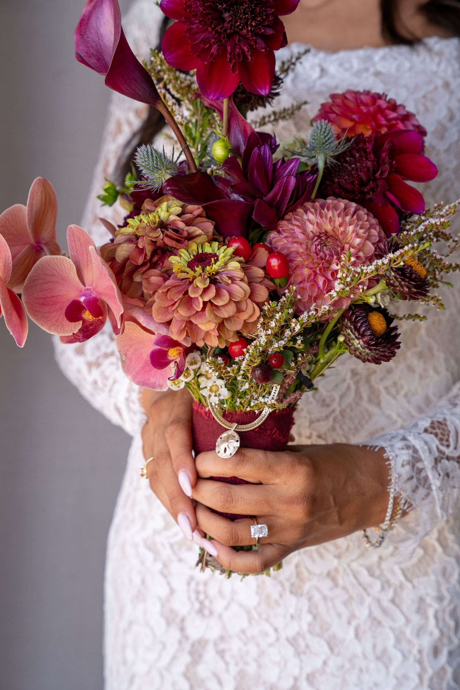 A vibrant wedding bouquet with burgundy and pink flowers held by hands wearing a lace sleeve and diamond ring.