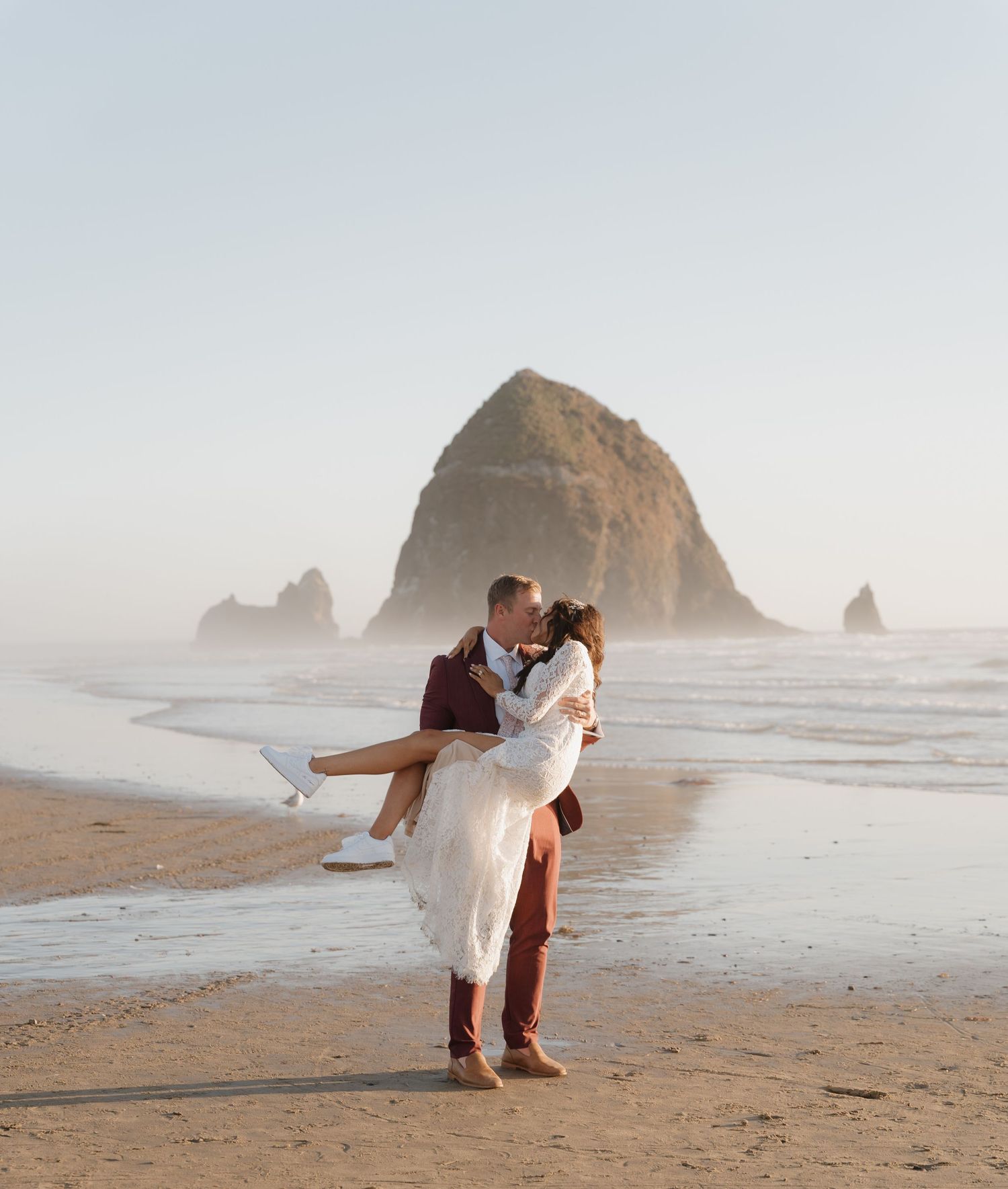 A couple embraces on a misty beach with Haystack Rock at Cannon Beach, Oregon in the background.