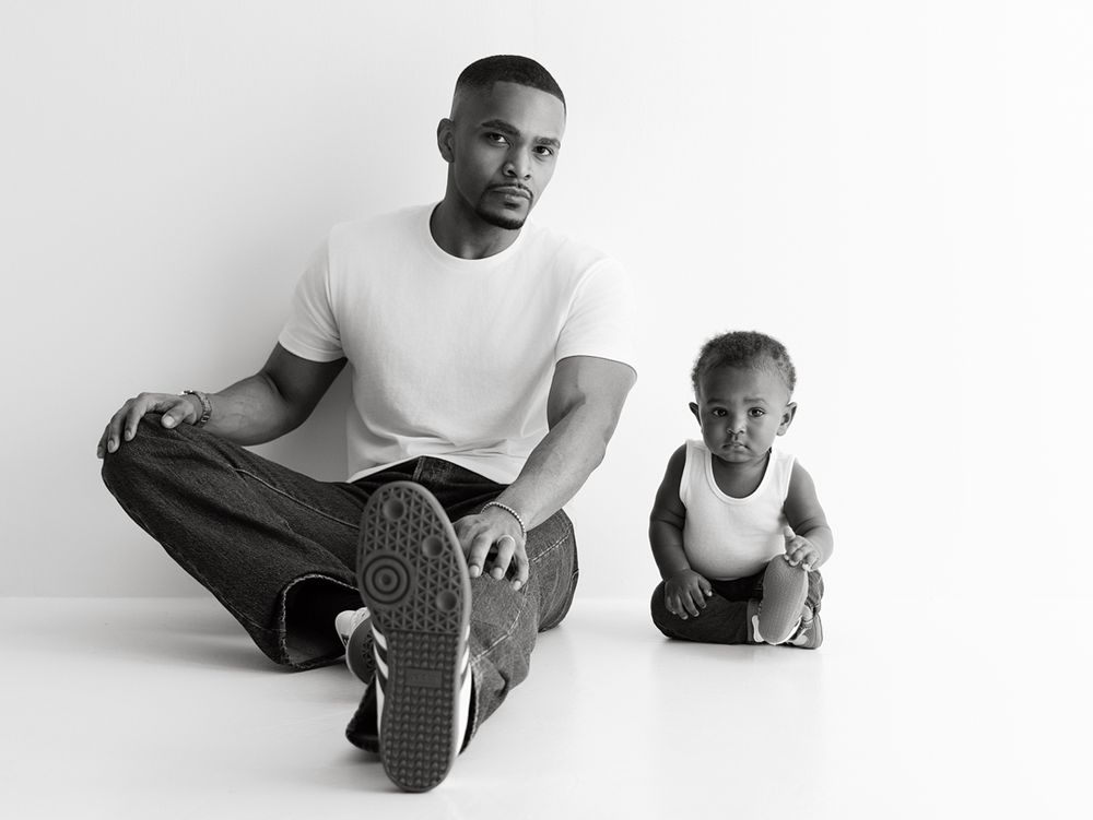 Black and white photo of a man in white t-shirt and jeans sitting with a toddler in a studio setting.