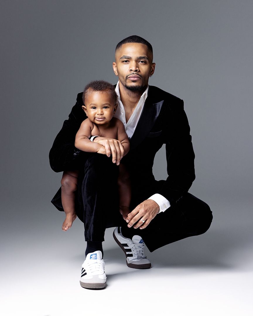 Man in black suit crouching while holding a baby, wearing Adidas sneakers, against a gray studio background.