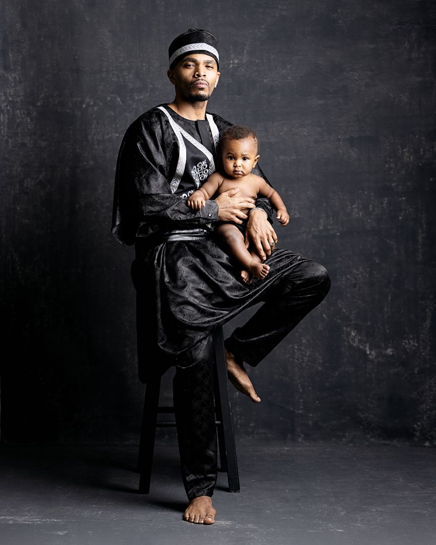 Man in black traditional African attire holds a baby while posing for a studio portrait photo.