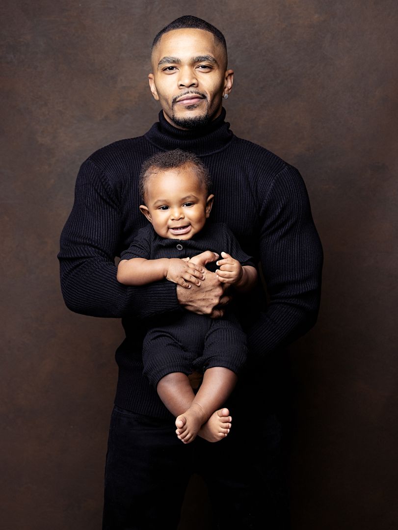 A muscular Black man in a black turtleneck holds a smiling baby dressed in black during a studio portrait session.