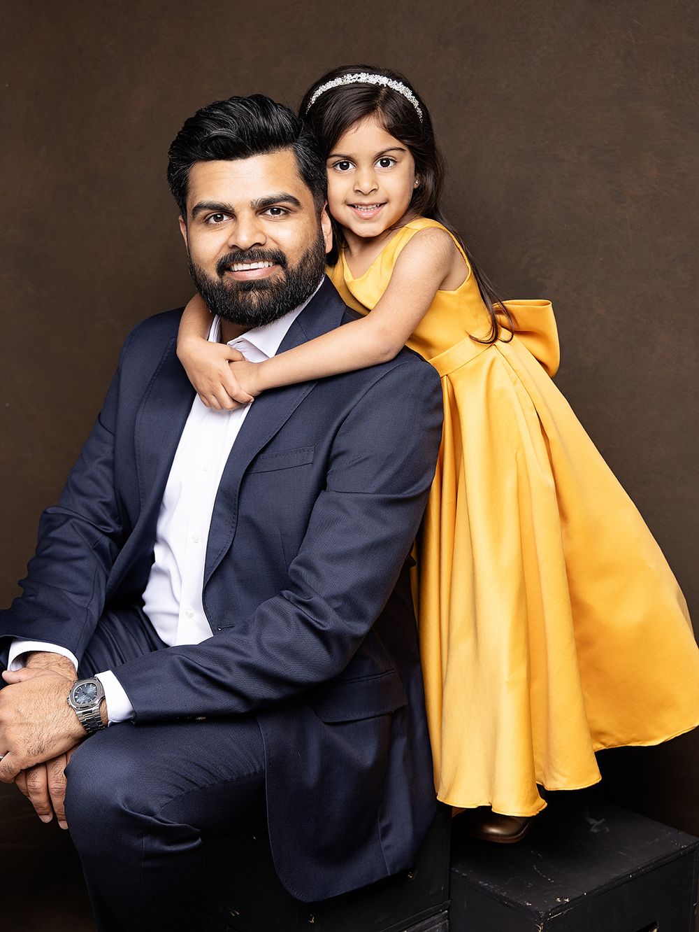 Portrait of an individual in a navy suit with a young child in a bright yellow dress posing together against a dark backdrop.