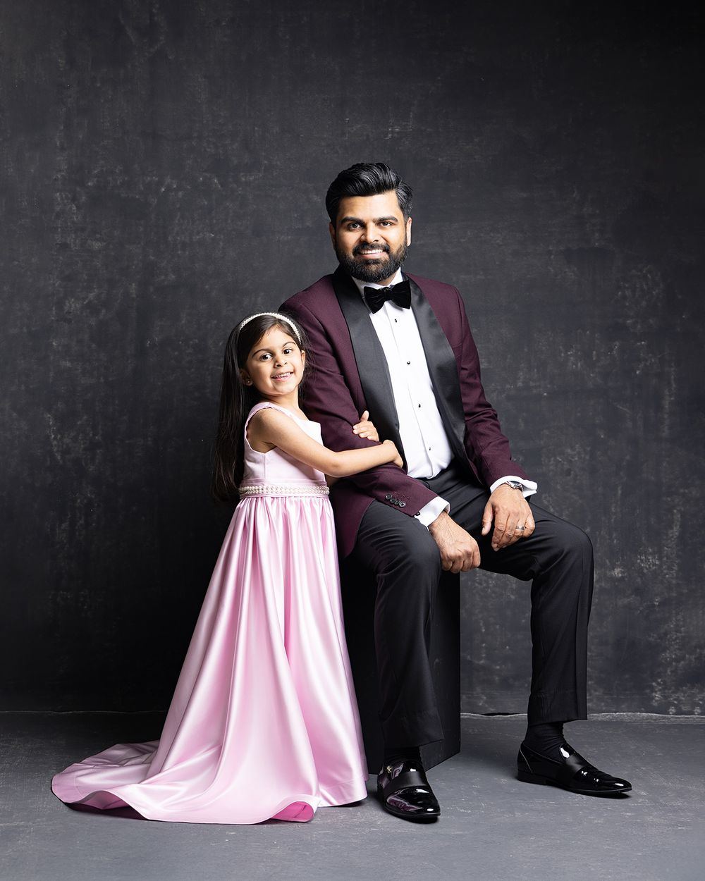 Formal portrait of two figures against dark backdrop - one in burgundy tuxedo seated, one in flowing pink gown standing.