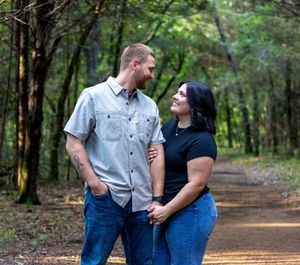 A couple shares a loving gaze while standing together on a wooded trail surrounded by lush green trees.