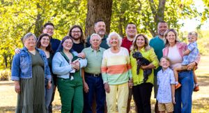 Large multi-generational family group poses together outdoors in casual clothing against autumn trees.