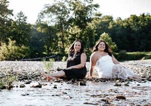 A sister photoshoot on a rocky riverbank during golden hour with a scenic natural backdrop.