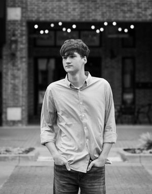 Young man in button-up shirt and jeans stands outdoors in front of brick building with string lights, black and white photo.