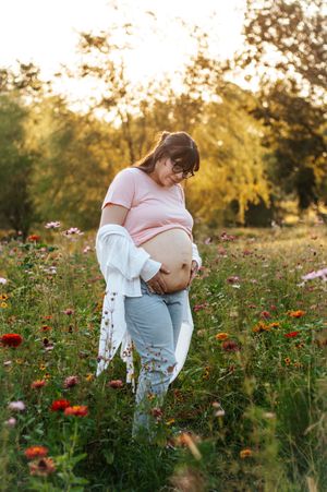 Pregnant woman in pink top and jeans cradling her baby bump in a wildflower meadow at golden hour.