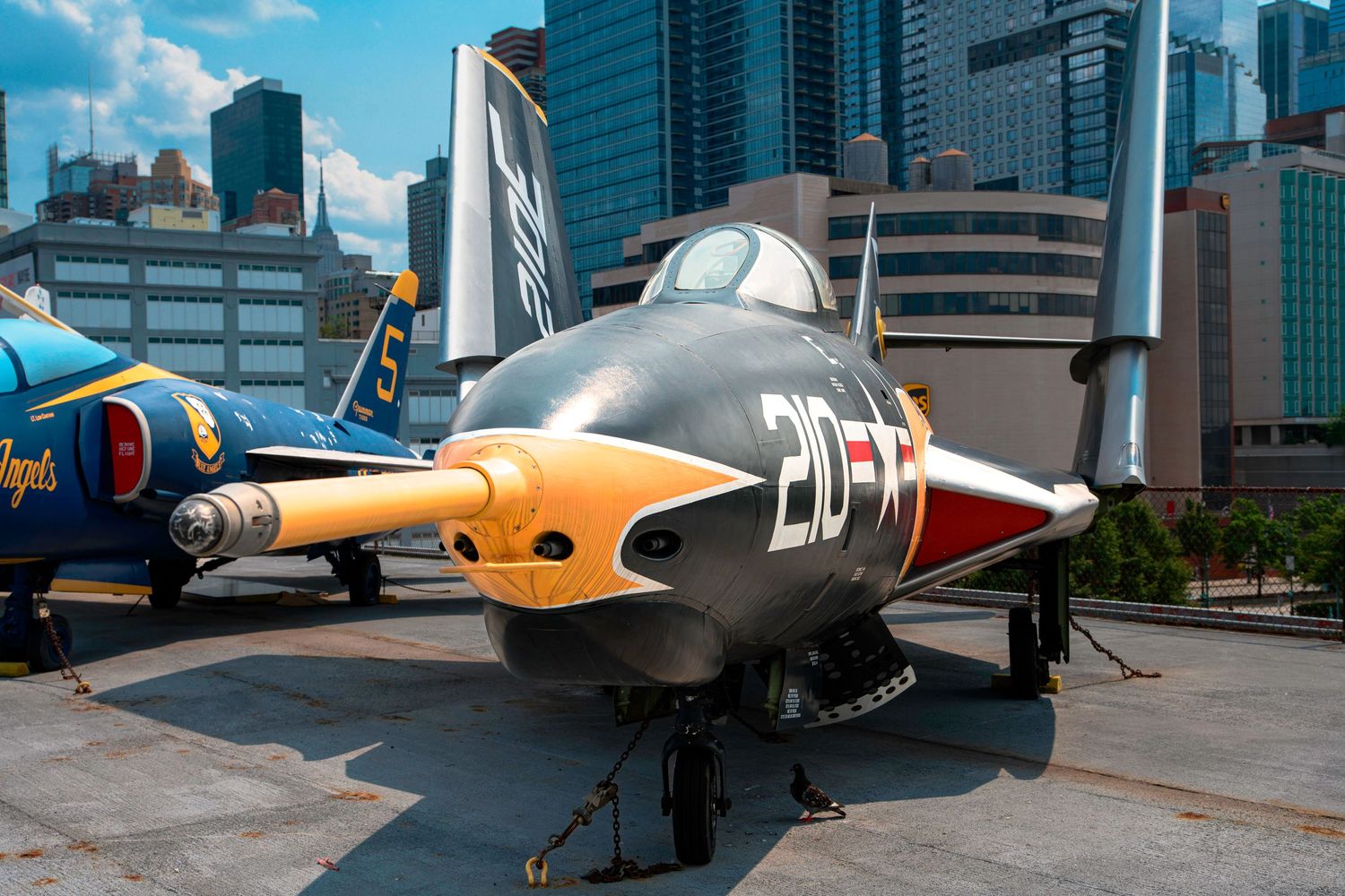 A yellow and red vintage military training aircraft displayed outdoors with modern buildings in background.