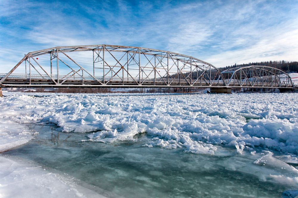 Chunks of ice float beneath a steel truss bridge on a partially frozen river under blue skies.