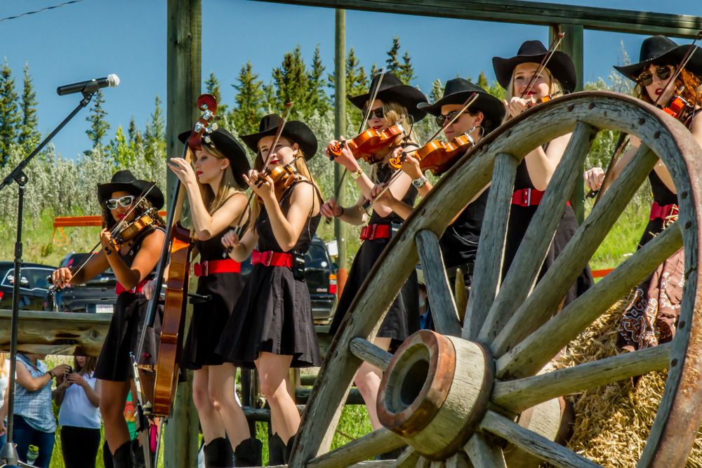 Young female violinists in black outfits performing outdoors near a rustic wagon wheel.