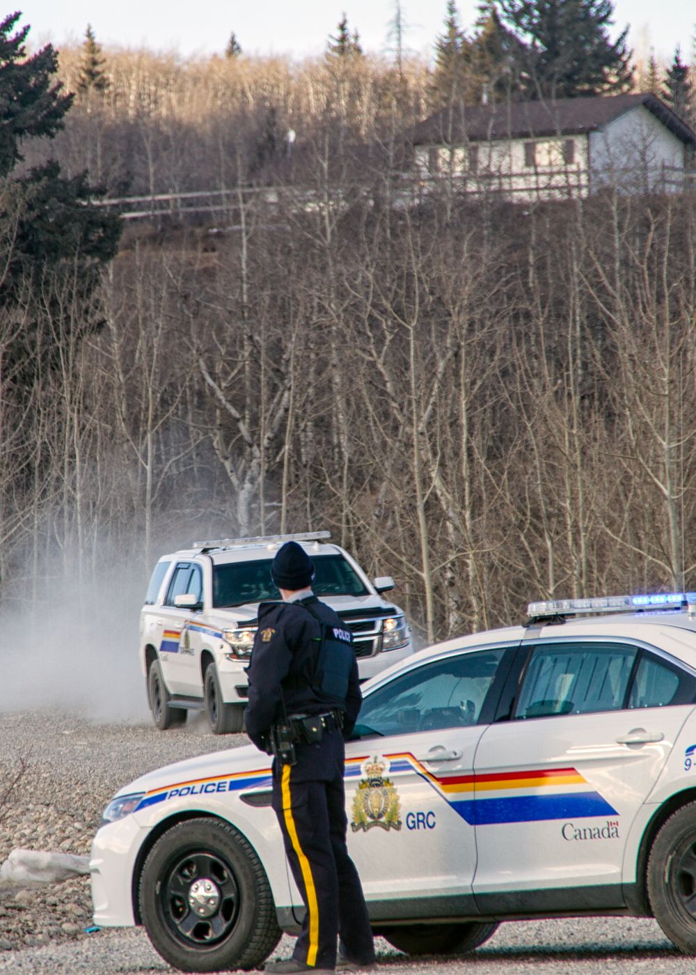 An RCMP officer stands beside a GRC police cruiser on a gravel road in a wooded area.