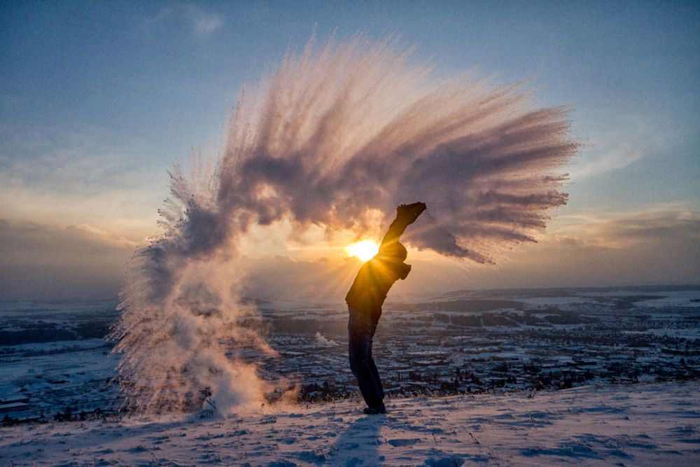 Person throwing boiling water into freezing cold air at sunset in snowy landscape.