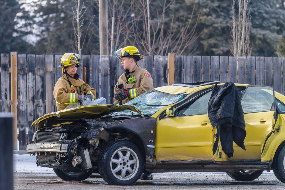 Two firefighters in yellow gear inspect a heavily damaged yellow car after a collision.