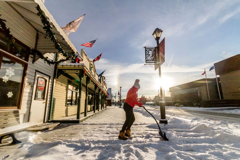 Person shoveling snow on a decorated small-town street at golden hour.