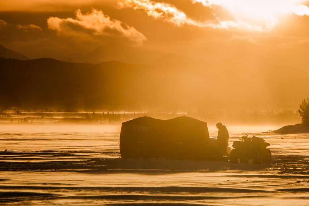 A silhouetted ice fishing hut glows against a stunning golden sunrise over a frozen lake.