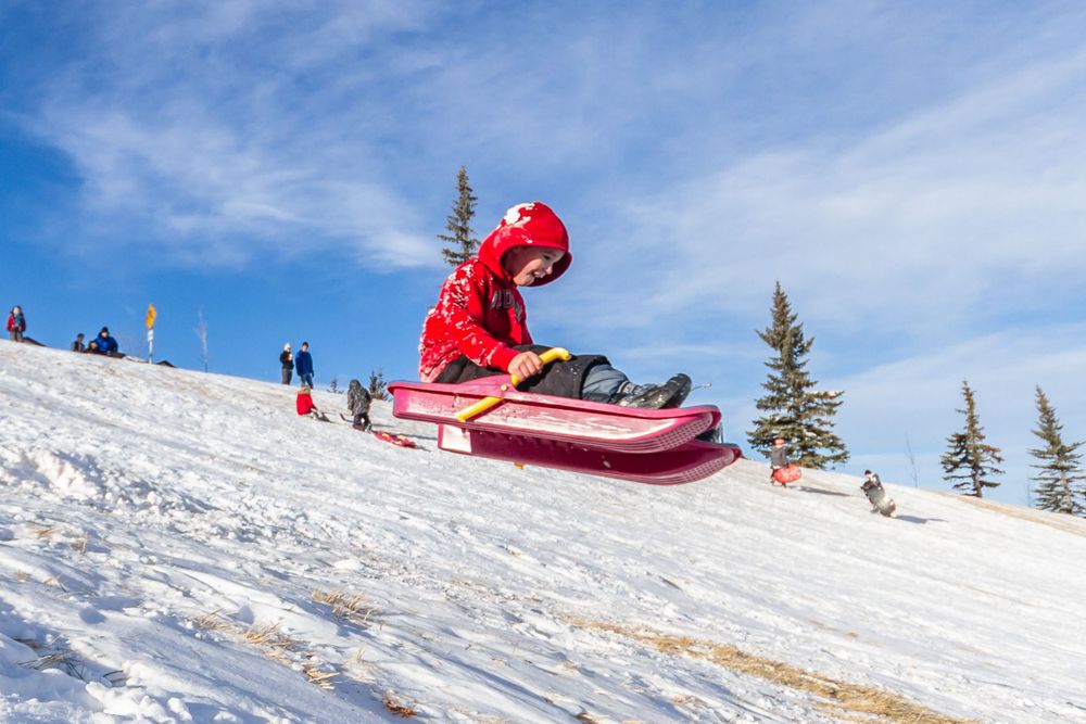 Child in red snowsuit sledding down a snowy hill on a bright winter day.