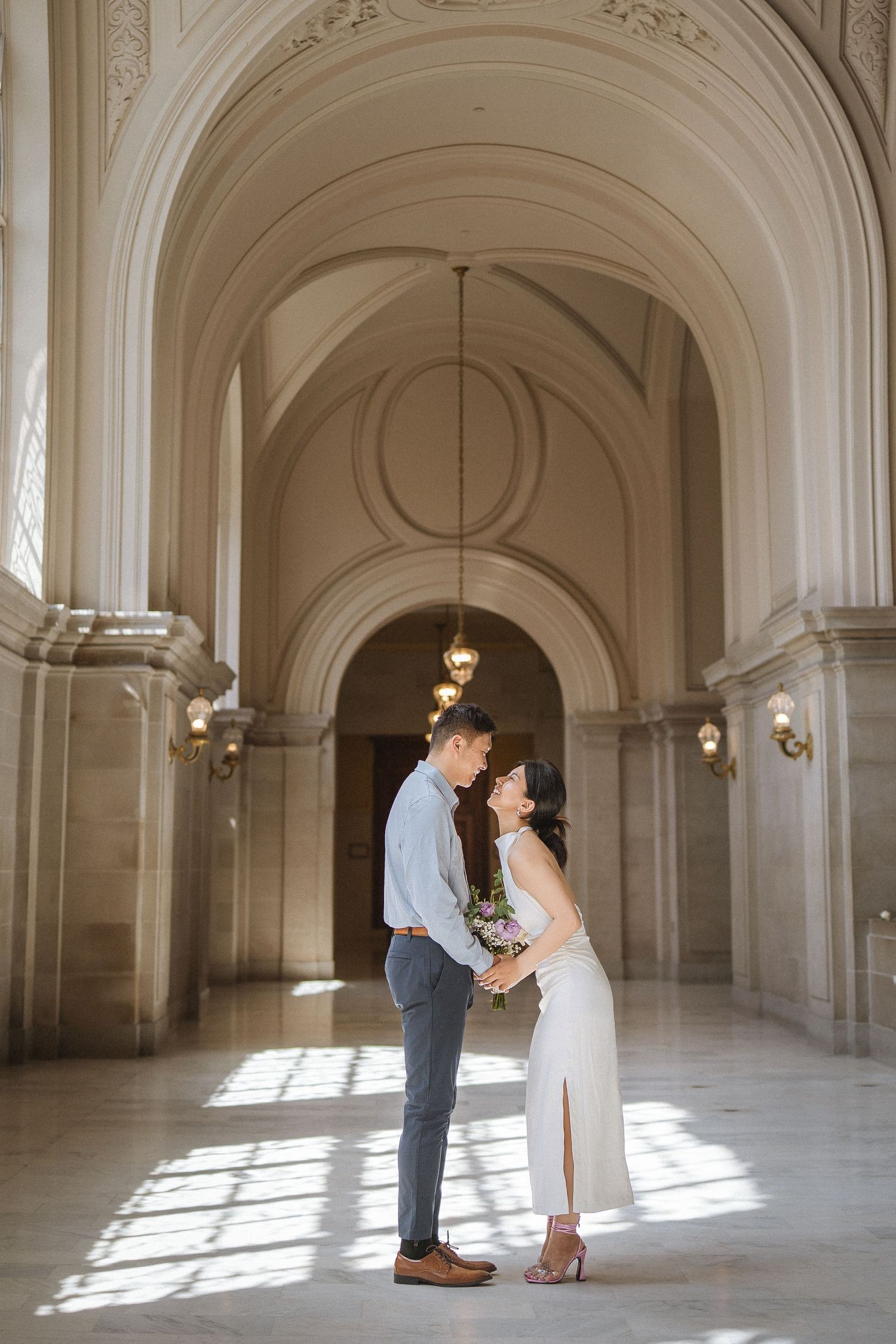 A couple dancing and twirling in an elegant white marble hallway with ornate architectural details and natural lighting.
