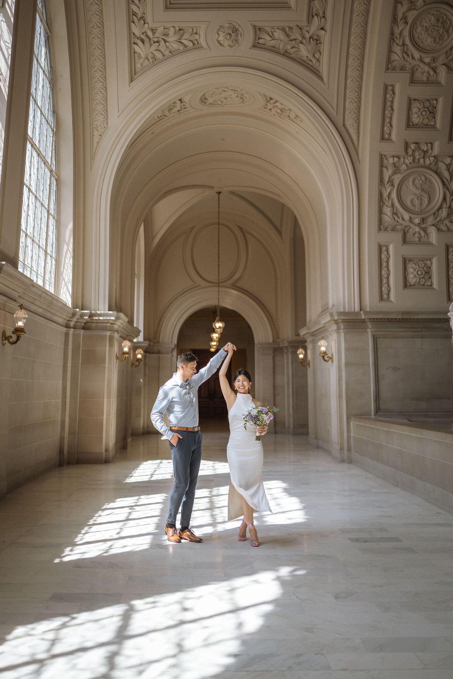 A couple poses playfully in a grand historic building with arched ceilings and streaming sunlight.