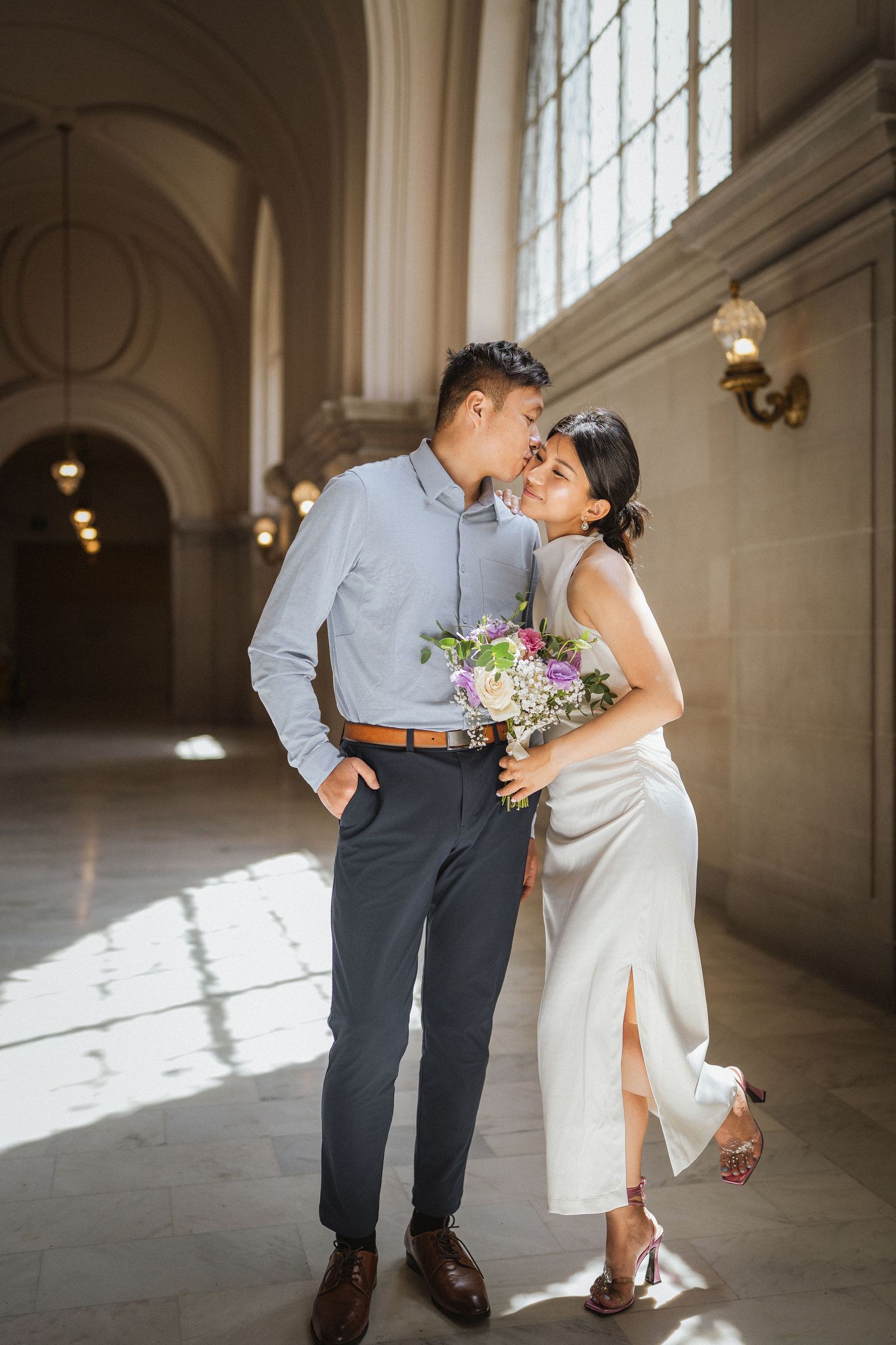 Wedding couple shares intimate dance in sunlit grand hallway with classic architecture.