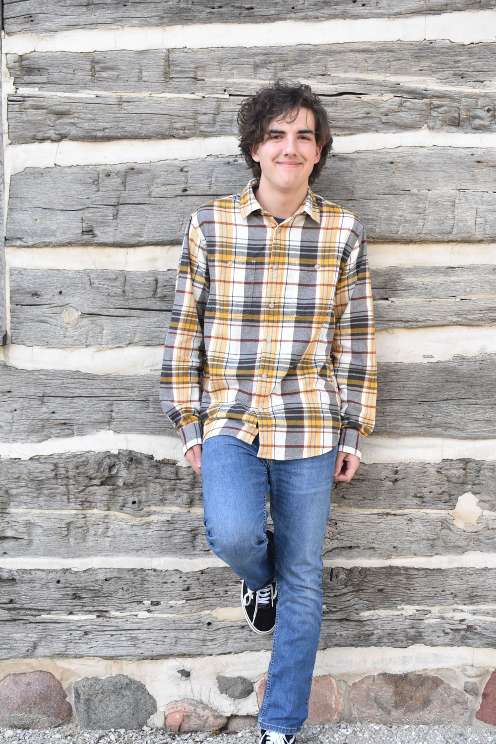Casual pose against rustic log cabin wall wearing yellow plaid shirt and jeans.