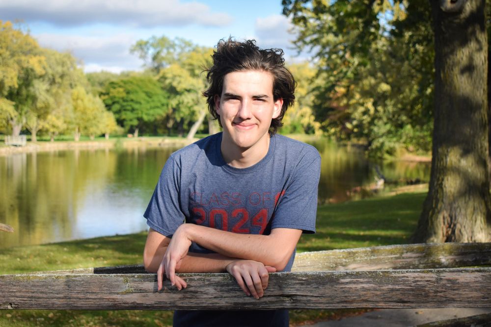 A person smiles while leaning on a wooden railing near a serene lake on a sunny autumn day.