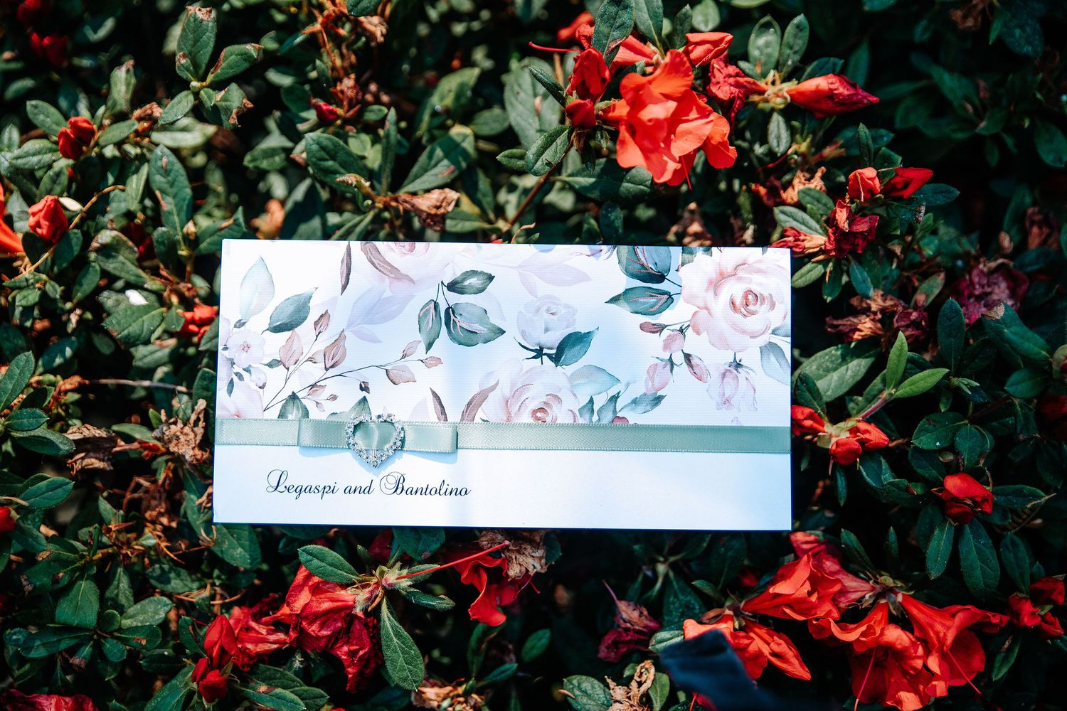 A blue and white floral wedding invitation placed among vibrant red flowers and green foliage.