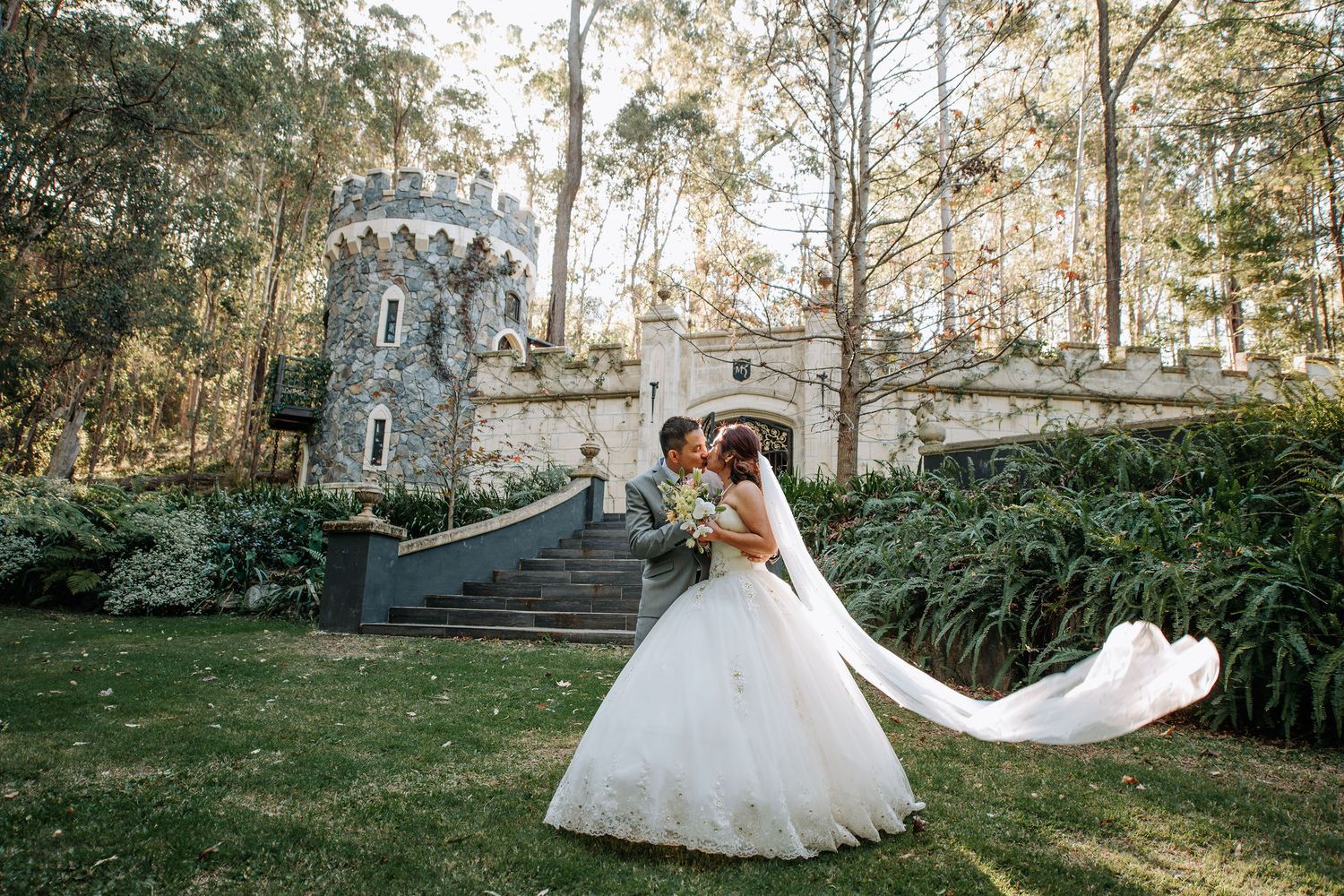 Bride in flowing white gown poses dramatically in front of castle-like stone structure.