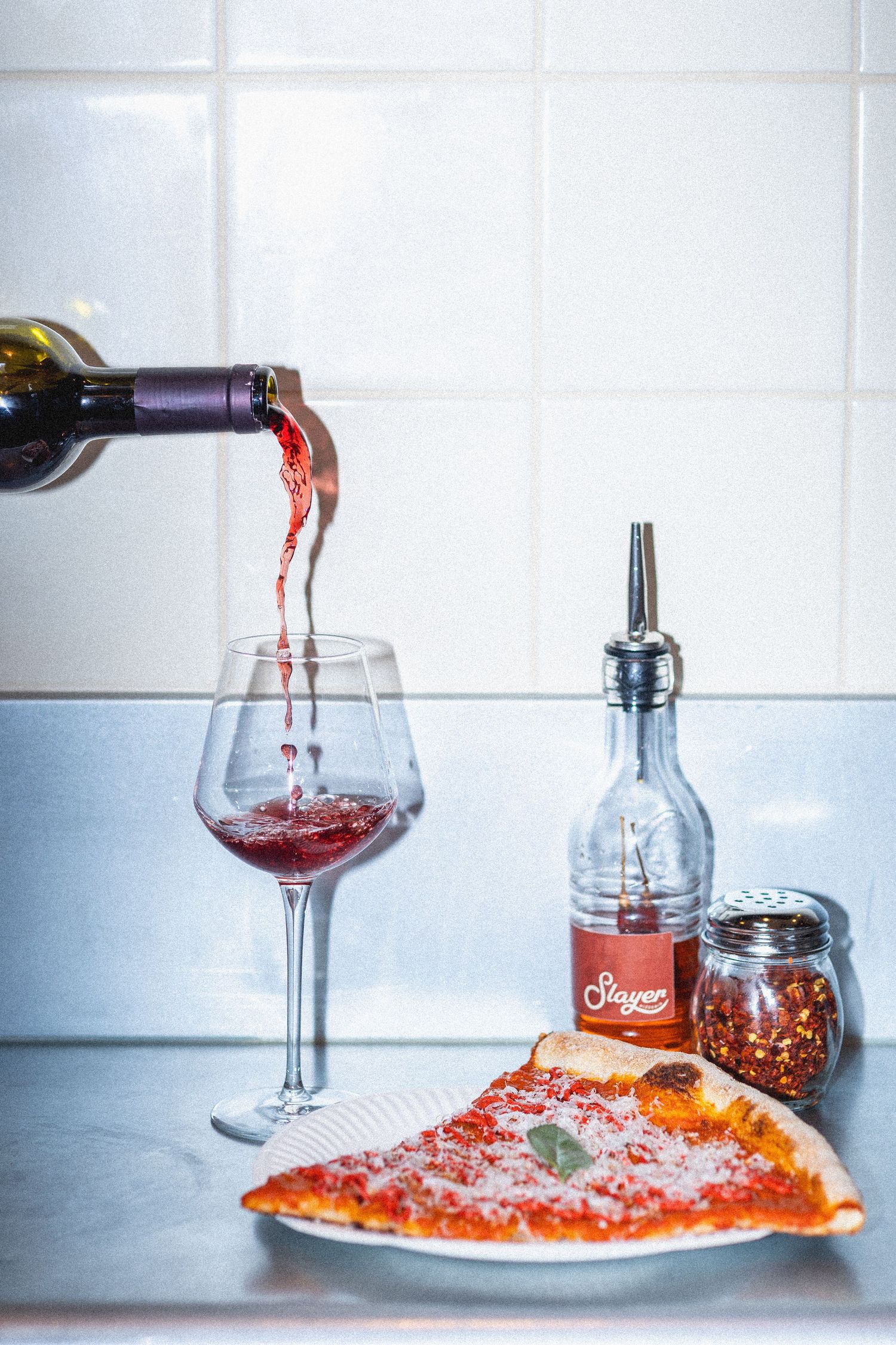 Red wine being poured into a glass beside a cheese pizza slice, olive oil, and spices on a counter.