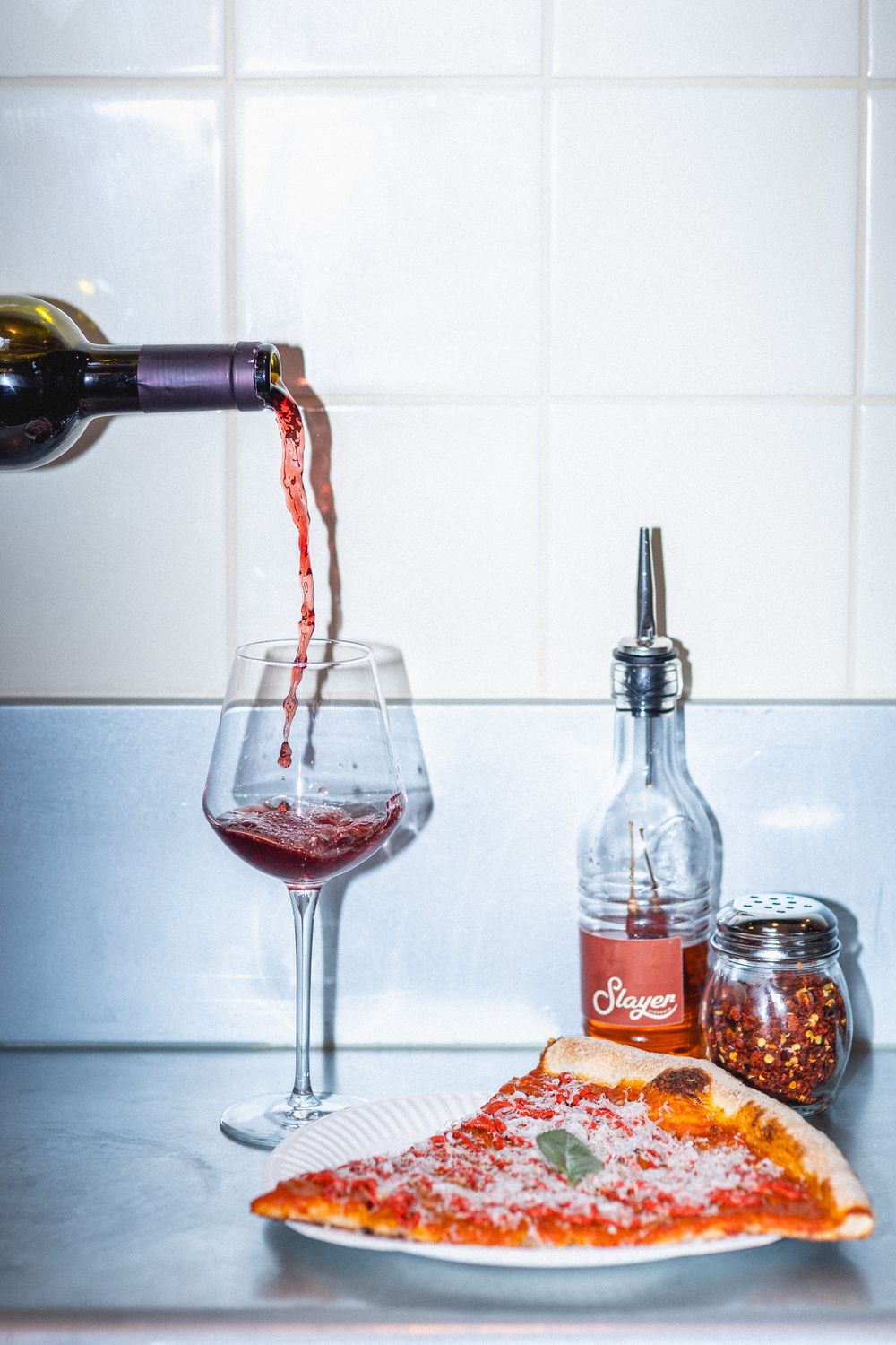 Red wine being poured into glasses next to a pizza on a white tiled background.
