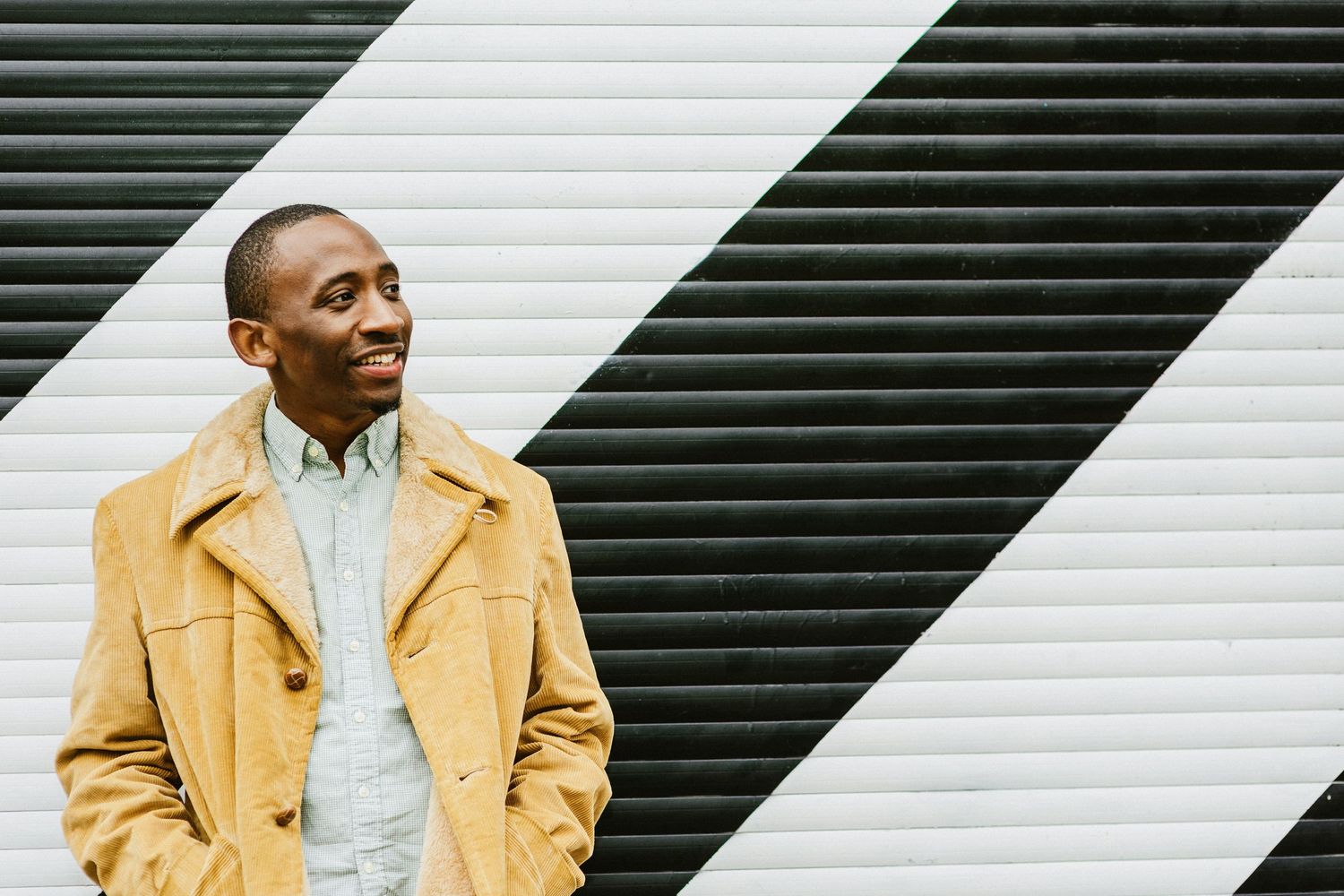 Smiling young Black man in yellow coat standing against black and white striped wall outdoors.