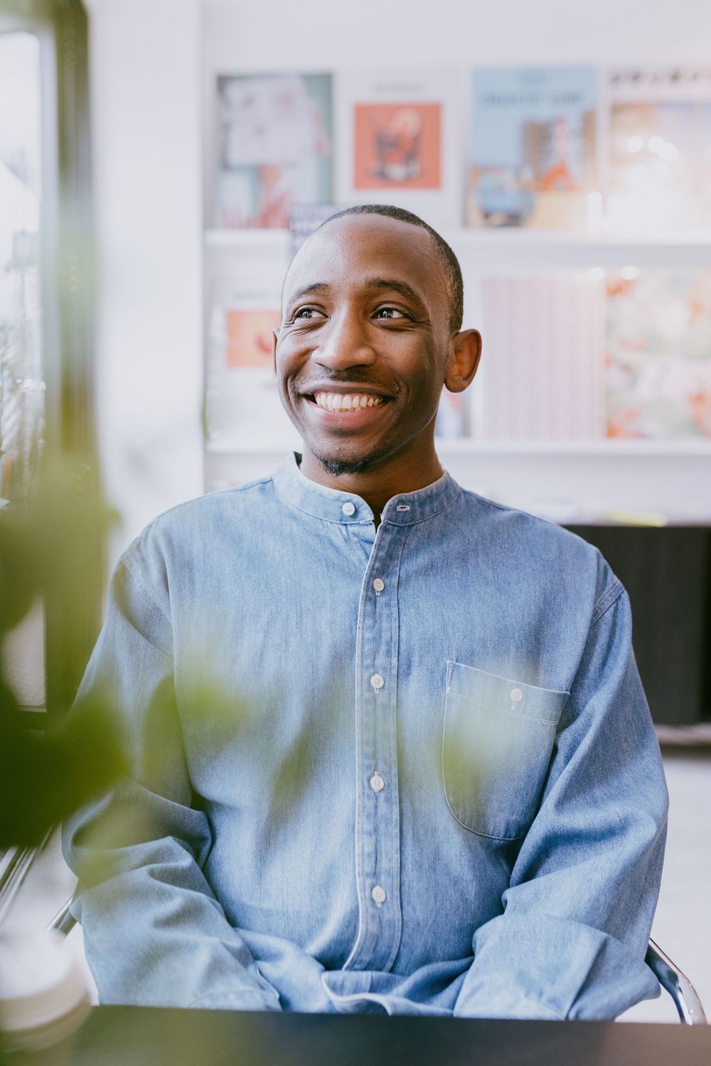 Smiling young Black man in a denim shirt sitting at a desk in a bright modern office space.