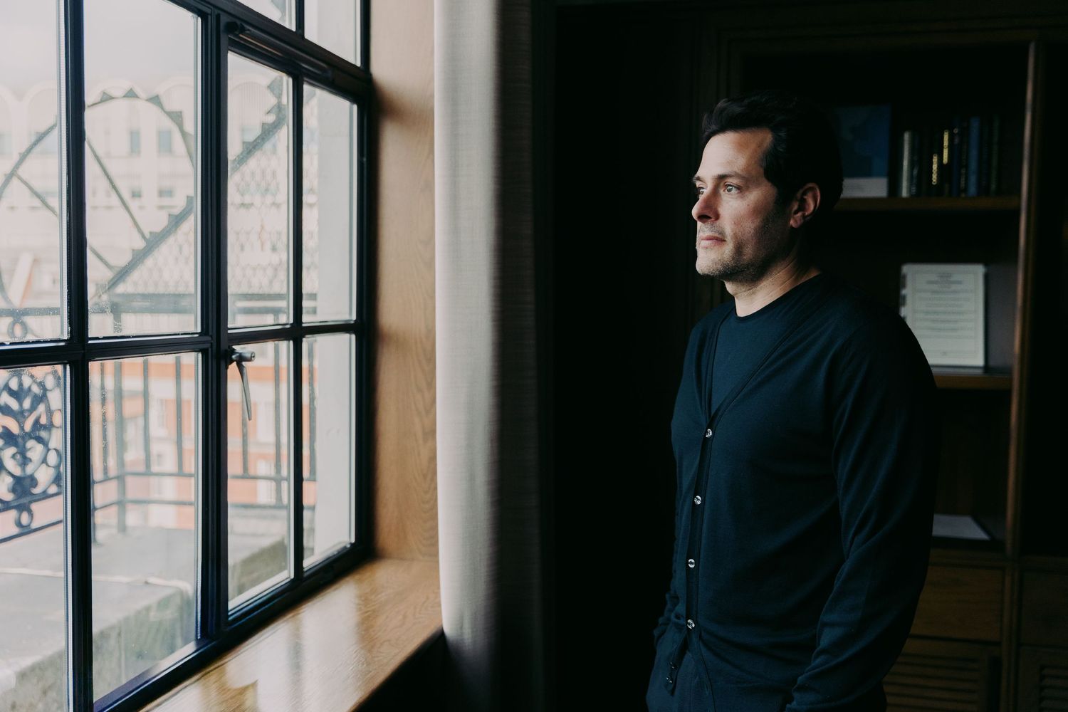 A thoughtful man in a dark blue shirt gazes out a large window in a dimly lit room with bookshelves.