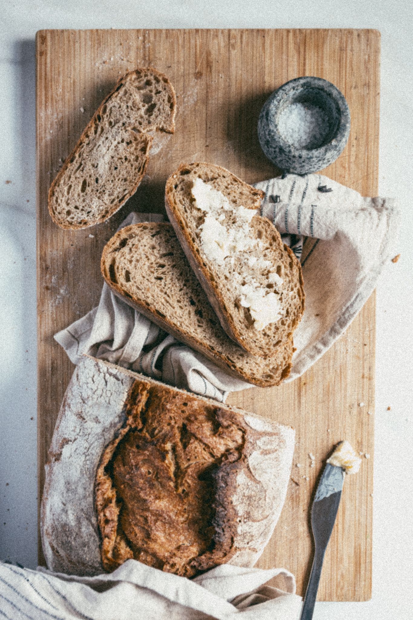 Rustic sourdough bread loaf sliced on wooden cutting board with linen cloth and mortar, overhead view.