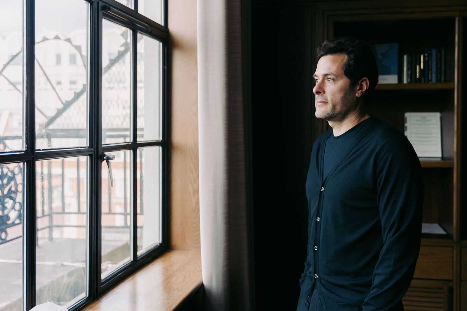 Person in black clothing looking contemplatively through large windows in an office setting.
